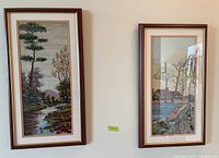 Photo shows two framed needlepoint art pieces hanging side by side on a wall, both with rural scenes and similar framing.