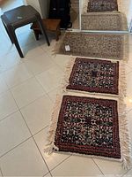Photo showing three rugs and two stools on a tiled floor, with two wool rugs having intricate patterns and fringe, one larger worn wool rug, one dark brown PVC stool, and one small wooden stool.