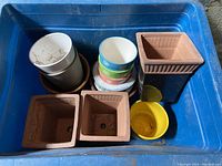 Top-down view of one bin showing two square brown clay pots, one dark blue rectangular pot, several stacked white and colorful circular ceramic pots, bright yellow pot, and dish plates underneath.