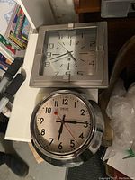 Photo of two clocks stacked on a surface. Top clock is square with a modern silver-tone frame. Bottom clock is a round Smiths clock with chrome casing.