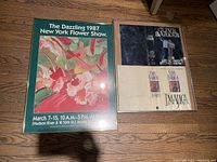 Photo of two framed pictures side by side on a carpeted floor. Left is a colorful floral poster for 1987 New York Flower Show in a gold frame. Right is a darker Clive Barker poster with visible text 'Clive Barker' and 'Imajica'.