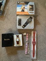Four watches laid out: Timex Ironman in packaging top, Bulova in box bottom left, pink girls watch bottom middle, red Swatch bottom right