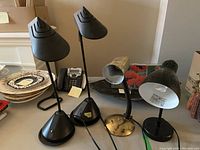 Photo of four table lamps in various sizes and styles shown on a surface. Two black modern lamps with conical shades and pyramid bases, one small brass gooseneck lamp, and one larger black adjustable lamp.