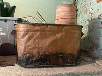 Sideline view of large rectangular antique tin container showing hammered texture with dark discoloration along the bottom edge. Container holds terracotta pots and plant wires inside.