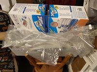 Two unopened boxes of Zip Lock Space Bag vacuum storage bags in blue and orange packaging with assorted loose clear plastic vacuum storage bags placed in a white bin on a wooden chair.