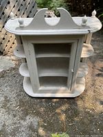 White wooden curio cabinet with glass door, decorative top trim, and side shelves shown outdoors.