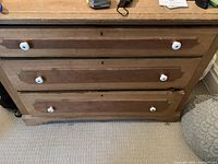 Front view of the light brown wooden dresser showing three drawers with white ceramic knobs and visible wear on the surface.