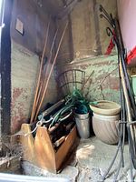 Photo showing a corner with stacked clay pot, metal pot, garden tools in wooden box, and shepherd hooks leaning against the wall.