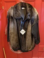 Front view of an otter fur jacket with leather sleeves and zipper closure hanging on a hanger against a red door.