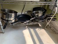 Shelf with assorted stainless steel pots and pans including saucepans with lids, frying pans and grill pan, all with black handles.