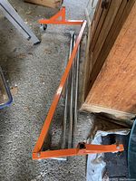 Orange triangular metal scaffolding frame pieces stacked with metal bars and wood planks underneath, showing wheels on one frame and clean wood surface.