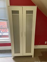 White wardrobe positioned in a corner against a red wall and near a window. It has two tall, narrow white doors with frosted glass panels and rectangular handles. The floor underneath is carpeted.