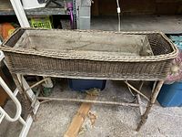 Photo of a large rectangular wicker planter on a wicker stand in a storage area showing natural wicker material and used condition.
