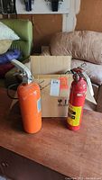 Two fire extinguishers, one red with a yellow label and one orange, positioned on a wooden table in front of a cardboard box.