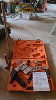 Paslode FSC200 stapler/nailer in orange plastic carrying case shown open on floor with manual and accessories visible.
