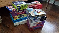 Photo showing stacked games named Scrabble, Reverse Charades, Balderdash, Wits & Wagers, Loaded Questions, Skip-Bo, with cribbage board and playing cards on top.