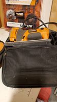 Full view of orange and black Rigid R3101 jig saw with carrying case and pack of extra blades in background.