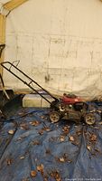 Full side view of the red gas lawn mower with black handle and wheels on a blue tarp with some leaves.
