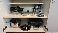 Photo of kitchen cabinet shelf holding assorted pots and pans including stainless steel and non-stick pans plus the electric pan.