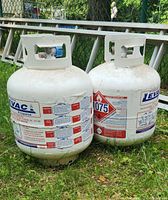 Two white Levac propane tanks, showing labels and hazard warnings, positioned on grass outdoors with a metal ladder in background.