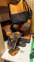 Four Rigid cordless drills in orange and black, charger, battery, and carrying bag on a white shelf with pegboard backdrop.