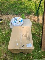 Photo of a closed cardboard box with 'Aqua Magic V' branding visible on the side and a coiled white and blue Aqua flex hose with brass connectors on top.