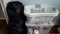 Black compressed North Face sleeping bag, white laundry basket on a plastic bin partially filled with yarn.