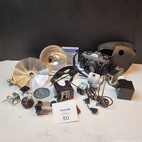 Photo showing vintage Pentax camera with lens, assorted flash bulbs, lens cleaning cloth, carrying case, power adapter, and other accessories all arranged on white surface with black background.