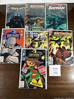 Photo showing front covers of 7 Batman comic books arranged on wood surface depicting various series and issue numbers.
