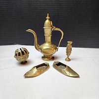 All five brass items displayed on a black background, showing the claw bell, pitcher, vase, and two shoe-shaped ashtrays with engraved feather patterns on the surface.