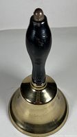 Front view of vintage brass school bell with black wooden handle on top, polished brass bell visible.