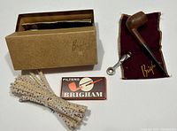 Full lot showing the wooden pipe in cloth pouch next to box of Brigham filters, metal tamper tool, pipe cleaners, and the original branded box.