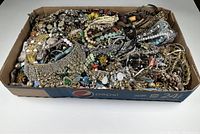 Wide view of large collection of various styles and colors of vintage estate jewelry in cardboard tray showing entire assortment, including chains, beads, and brooches.