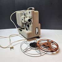 Side and rear view of the Eumig P8 projector showing lens, mechanical controls, electrical cord, and one reel loaded with another beside it.