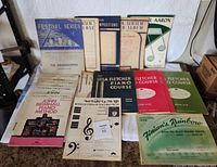 Photo showing the full assortment of 16 vintage piano music and teaching books laid out on a table, including titles such as 'Festival Series The Grasshopper,' 'Leila Fletcher Piano Course,' 'John Brimhall Piano Method,' and 'Finiario's Rainbow Easy-To-Play Piano Solos'. Covers exhibit classic mid-20th-century style graphics and typography.