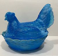 Side view of a bright blue slag glass candy dish shaped like a hen sitting on a basket weave patterned nest with visible texture and marbled white streaks.