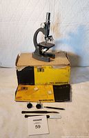 Scene with vintage Tower 750X microscope on top of worn original box, accompanied by manual, two lenses, tweezers, spatula, and scalpel tool.
