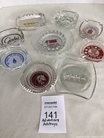Overview of nine different advertising ashtrays, various shapes and logos visible on a white background.