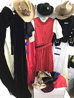 Full lot view showing hanging costumes (black hooded velvet cape, red dress with white blouse, clown costume, Bavarian dress), child's velvet jacket on chair, and hats including straw hat with floral decoration, green felt hat, and beige felt hat with braided band.