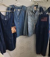 Photo shows four denim clothing pieces hanging: sleeveless jumper dress, embellished shirt, distressed shirt, and capri overalls.