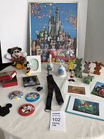 Photo showing full group of Disney memorabilia items including plush toy, framed poster, bells, cards, badges, and various figurines and collectibles on a table.