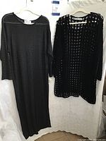 Two black cover ups hung on white hangers against white background, one floor length and mesh knit, the other shorter and windowpane style knit.