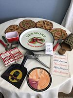 Wide view of all vintage kitchen items on white cloth showing Pyrex dish, utensils, Vermont tray, trivets, recipe booklet, and metal tools
