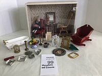 Photo shows the overall layout of the miniature diorama box with floral wallpaper, red cradle, rocking chair, clawfoot tub, birdcage, candlesticks, and various small decorative items spread around a white surface.