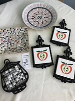Photo showing all six trivets including pastel round mosaic plate, multicolor square mosaic plate, and four black metal trivets with tile inserts.