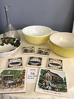 Photo of two yellow Pyrex mixing bowls, clear decanter, ceramic strawberry strainer, and eight decorative trivets arrayed on table.