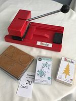 Photo showing red Sizzix manual die cutting machine with handle, wooden-framed geometric shapes die, and two packaged dies (snowflake and Christmas tree).