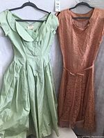 Two full dresses on hangers side by side, showing mint green dress with full skirt and apricot orange lace dress with belt