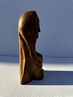 Side view of the wooden sculpture emphasizing the profile of the woman and the detailed carving of the hood and clothing.
