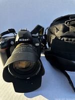 Front view of Nikon D70 camera with zoom lens and lens hood attached, next to Nikon branded carrying case.